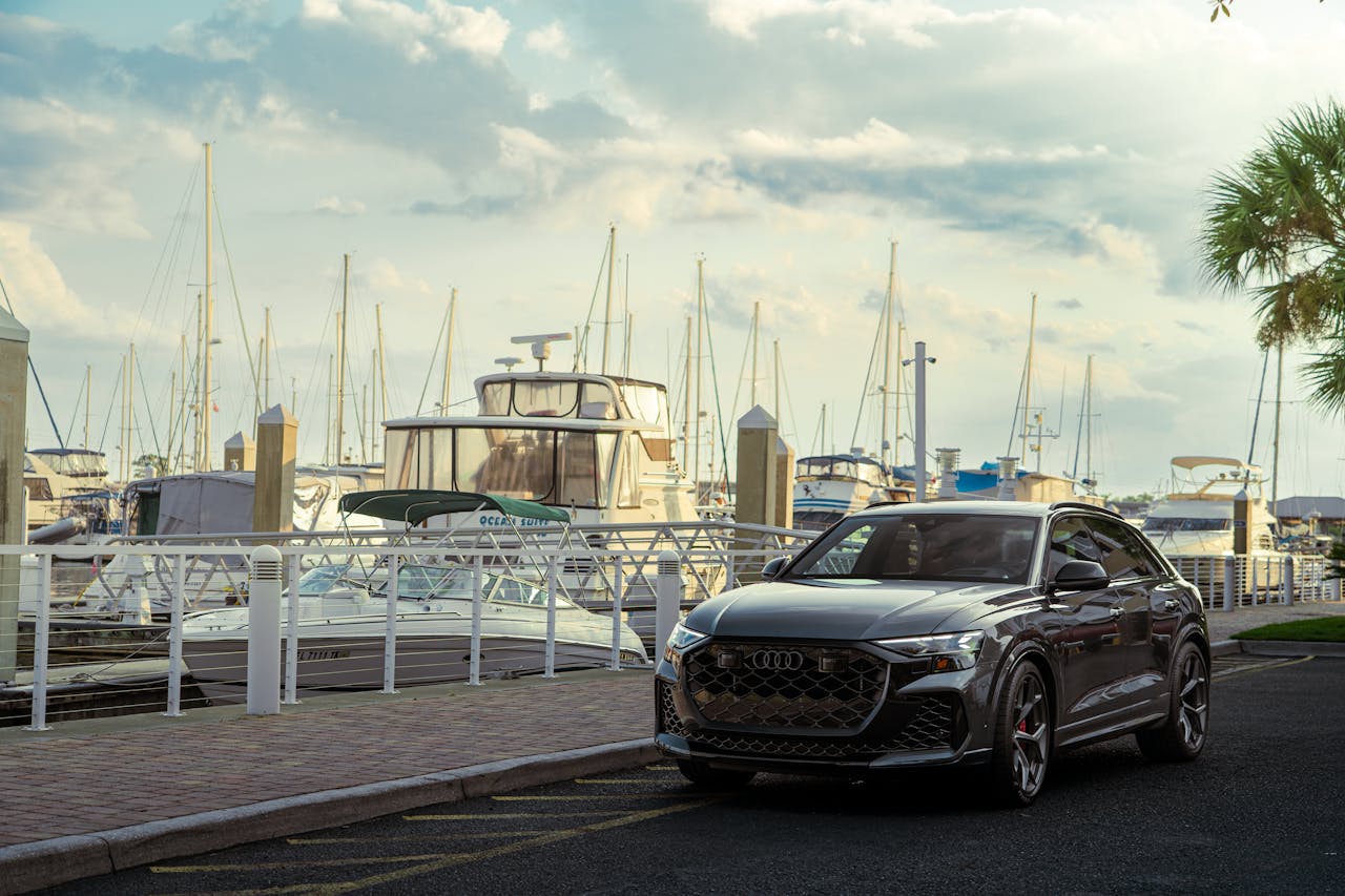 Sleek luxury car parked beside yachts in a scenic marina setting under a cloudy sky.